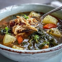 A steaming bowl of Collard Greens, Chicken and Vegetable Soup garnished with fresh herbs and a lemon wedge, served beside crusty bread. 